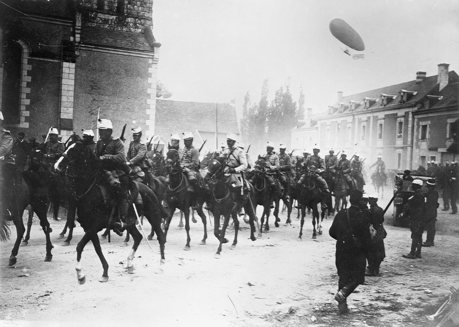 Chasseurs à cheval parade through town while a dirigible flies overhead in 1914.